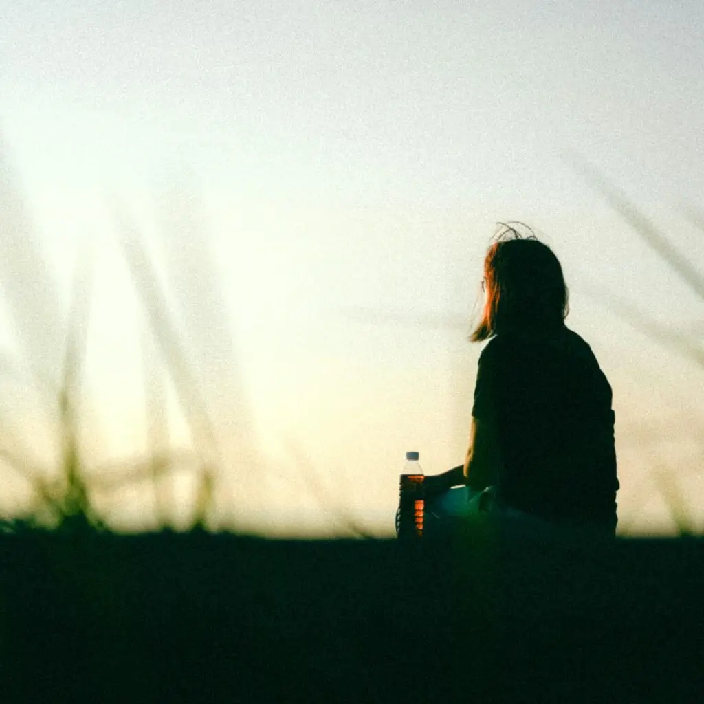 A woman sitting outdoors at sunset holding a bottle, captured in a peaceful, contemplative moment.
