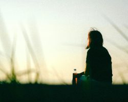 A woman sitting outdoors at sunset holding a bottle, captured in a peaceful, contemplative moment.